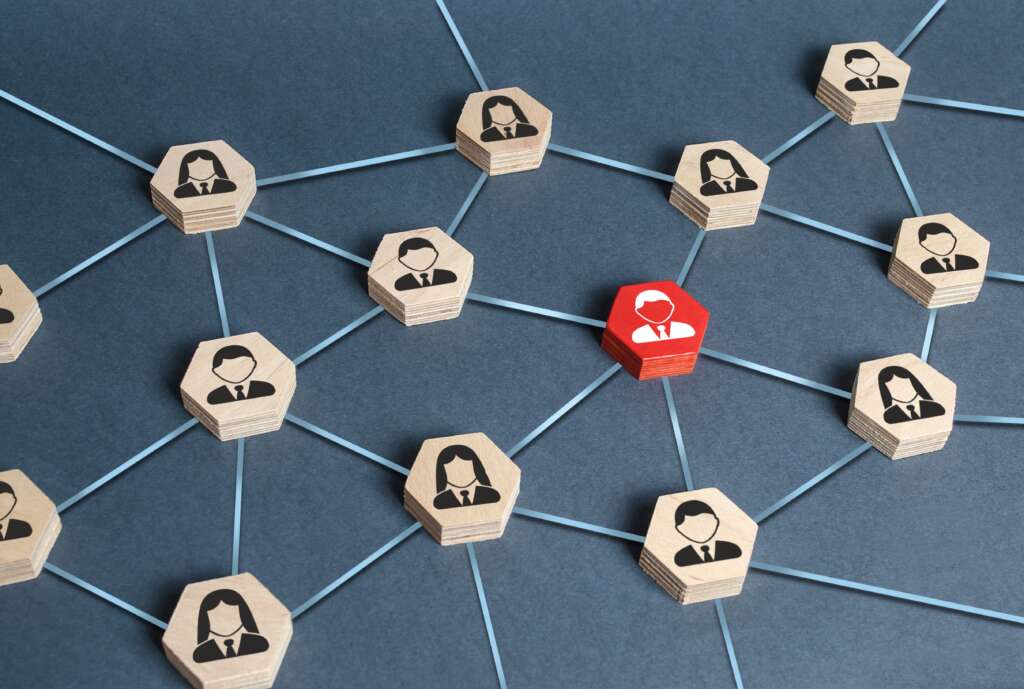 A conceptual image showing a network of wooden hexagonal tiles with illustrations of business people in suits, connected by blue lines. One tile is highlighted in red, representing a central figure in the network.