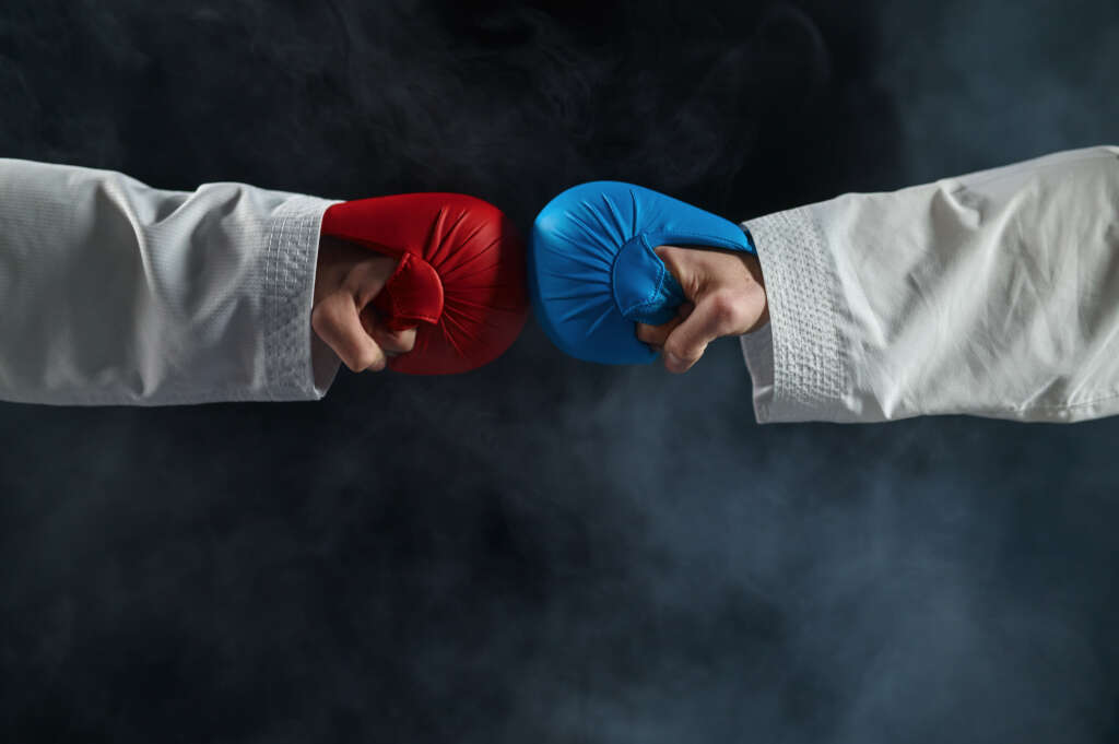 Two martial artists in white uniforms engaging in a friendly sparring match, each wearing a red and blue glove, with their fists gently touching in the center. The background features a smoky, dramatic effect.